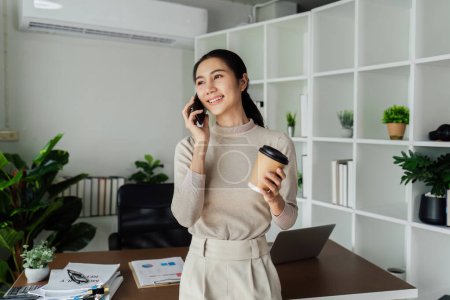 Geschäftsfrau hält eine umweltfreundliche Kaffeetasse aus Papier in der Hand und spricht mit dem Handy in netter Büroumgebung.
