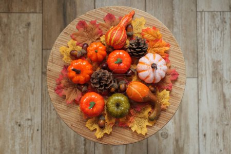 Photo for An arrangement of of plastic pumpkins, acorns, pinecones, and autumnal fabric leaves, set on a circular disc of wood, above ceramic wood effect tiles. - Royalty Free Image