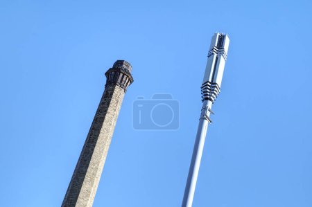 Photo for Bradford, UK 11 11 2023 - A  telecommunications mast is seen beside a victorian mill chimney on the outskirts of Bradford. Shot at an angle to make the image more dramatic and quirky - Royalty Free Image
