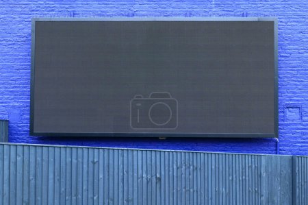 LED advertising hoarding wall mounted outdoors on a blue brick wall with blue fence panels in front. The hoarding appears off, or may only be displaying black.