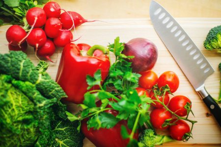 Composition culinaire : Gros plan du couteau du chef sur une planche à découper en bois avec légumes assortis - brocoli, tomates, oignons et poivrons (Image tonique, Vue du dessus, Image 4K)