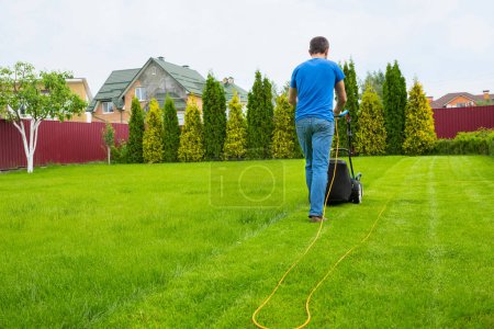 Téléchargez les photos : Un jardinier avec une tondeuse à gazon coupe l'herbe dans le jardin dans la cour arrière de la maison. - en image libre de droit