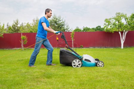 A gardener with a lawn mower is cutting green grass in the garden in the house backyard.