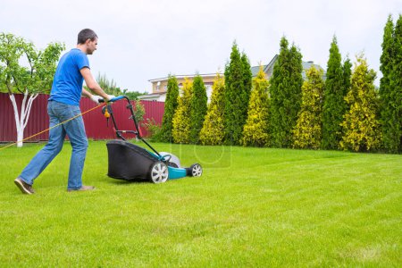 Téléchargez les photos : Un travailleur dans le jardin avec une tondeuse à gazon coupe de l'herbe dans une journée ensoleillée d'été dans la cour arrière. - en image libre de droit