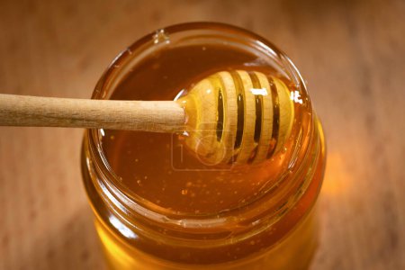 A glass jar of fragrant honey and a wooden honey dipper on a wooden background, close-up.