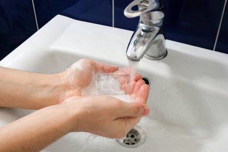 Foto de Manos masculinas en jabón, sobre un grifo con agua en el baño. Concepto de higiene. - Imagen libre de derechos