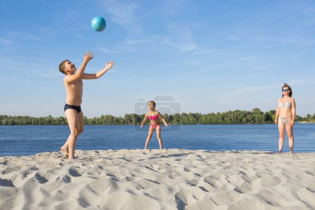 Téléchargez les photos : Les enfants heureux jouent avec le ballon avec leurs parents sur la rive de la rivière, pendant la saison estivale. Loisirs en famille sur l'eau. - en image libre de droit