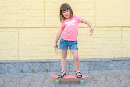Téléchargez les photos : Élégante, petite fille avec une planche à roulettes sur un fond de mur jaune, posant pour la caméra. - en image libre de droit