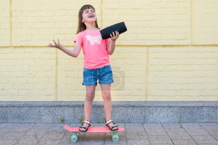 Téléchargez les photos : Enfant à la mode écoutant de la musique debout près d'un skateboard, sur fond de mur de briques jaunes. - en image libre de droit