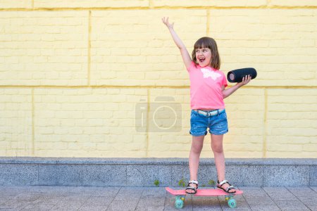 Téléchargez les photos : Jolie petite fille avec une planche à roulettes écoutant de la musique sur un fond de mur de briques jaunes. Enfant, hipster. - en image libre de droit