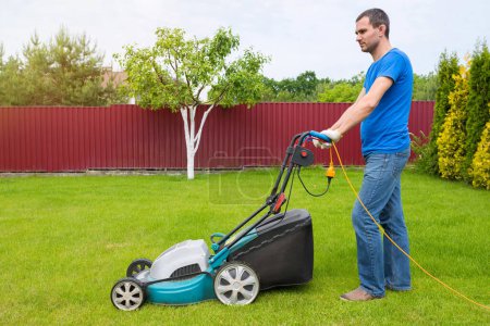 Téléchargez les photos : Travailleur avec une tondeuse à gazon tondre l'herbe dans le jardin. Vue latérale. - en image libre de droit
