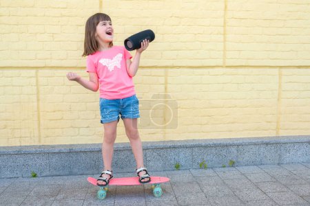 Téléchargez les photos : Petite fille élégante écoutant de la musique debout sur un skateboard, sur le fond d'un mur de briques jaunes. Style de vie. - en image libre de droit