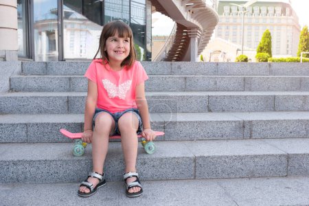 Téléchargez les photos : Petite fille à la mode est assis sur un skateboard dans la ville. Style de vie. - en image libre de droit