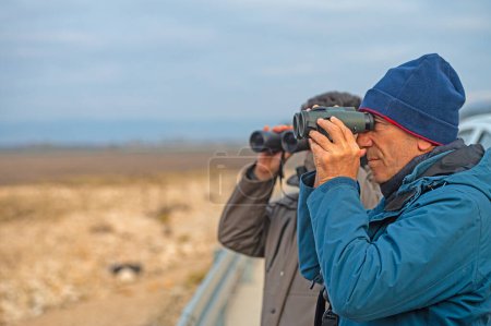 Foto de Un hombre vistiendo un abrigo azul y una boina está observando aves con prismáticos en la mano. - Imagen libre de derechos