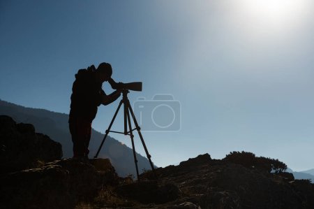 Silueta de un hombre observando aves con un telescopio en un trípode junto a un lago.