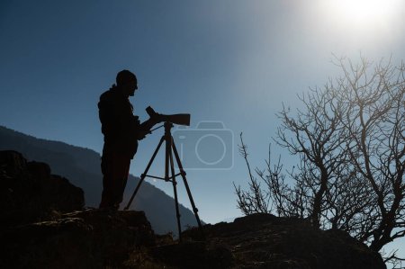 Silueta de un hombre observando aves con un telescopio en un trípode junto a un lago.