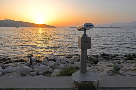 rear view closeup of coin operated metallic binoculars on concrete pier at the sea shore with sunset sky in the background