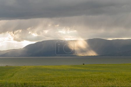 Green crop field by Lake Burdur, mountains and a beam of light hitting the earth in cloudy weather.