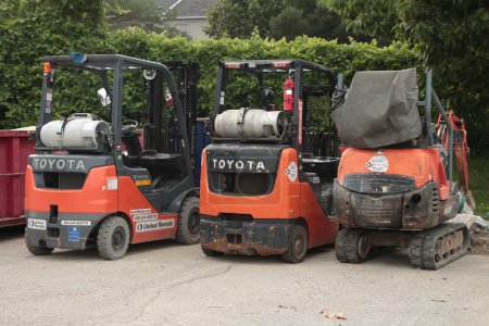 Photo for Two toyota propane forklifts orange and black parked next to another orange piece of machinery in a parking lot next to a dumpster and with bushes behind - Royalty Free Image