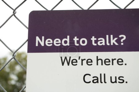 need to talk were here call us sign in white and black print on purple and white background, fastened to fence with blurred tree and sky in background