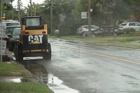 Foto de Bobcat aparcado al lado de la carretera de lluvia húmeda con la persona sentada en él visto desde atrás, charcos camino mojado - Imagen libre de derechos