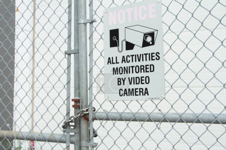 Photo for All activities monitored by video camera white rectangle sign with black print and illustration picture of video camera against silver diamond fence - Royalty Free Image