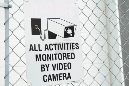 Photo for All activities monitored by video camera white rectangle sign with black print and illustration picture of video camera against silver diamond fence, close up - Royalty Free Image