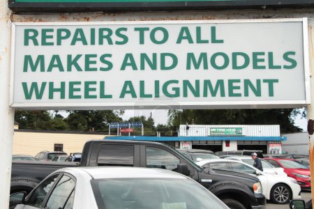 Photo for Mechanic shop sign with busy parking lot, sign says repairs to all makes and models wheel alignment in green writing on white background top frame, person in parking lot - Royalty Free Image