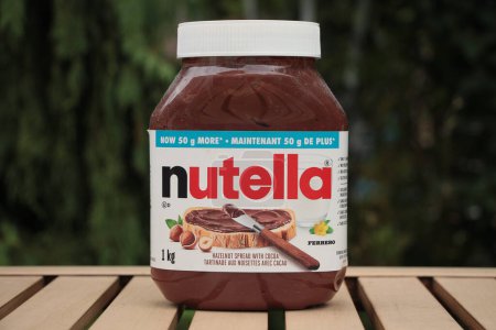 Foto de Nutella avellana untada con contenedor de cacao al aire libre en la mesa marrón claro con verde detrás, de cerca - Imagen libre de derechos