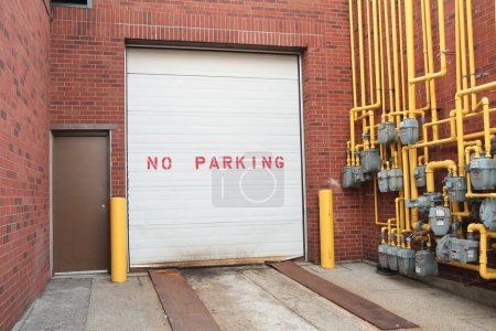 Foto de Sin texto de escritura de estacionamiento en la puerta del garaje blanco junto a la puerta de metal marrón en la pared de ladrillo y medidores de gas con tuberías amarillas - Imagen libre de derechos