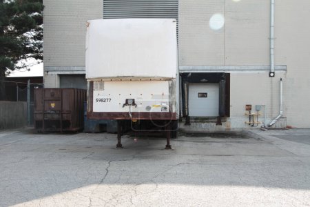 Photo for Two receiving bay garage doors next to each other in summer with one closed and the other with a large stationed parked white truck trailer storage in it, shot straight on - Royalty Free Image