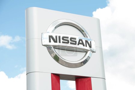 Photo for Nissan silver logo sign exterior outside outdoors on white red rectangle pillar with space in between with clouds sky behind with sun coming out behind clouds and shining bright on sign, close up - Royalty Free Image