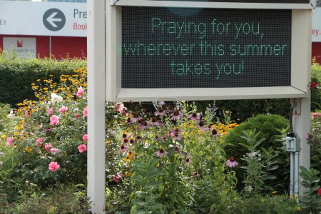 Téléchargez les photos : Signe électronique numérique électrique d'église dans le jardin de plante de fleur qui dit priant pour vous partout où cet été vous emmène - en image libre de droit