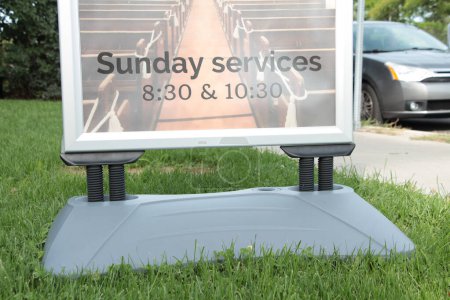 Photo for Sunday services 830 and 1030 time sign information on grass lawn next to sidewalk and street road in summer on bright day with car vehicle parked on road street - Royalty Free Image