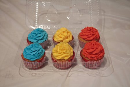 Photo for Six cupcakes in plastic container holder on table with white tablecloth with blue yellow and red icing decoration on top - Royalty Free Image