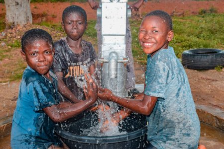 Foto de Abuja, Nigeria - 02 de agosto de 2021: Niños africanos se divierten mientras expresan felicidad y risa mientras juegan con agua limpia bajo el sol en una comunidad rural. Niños alegres y agradecidos - Imagen libre de derechos