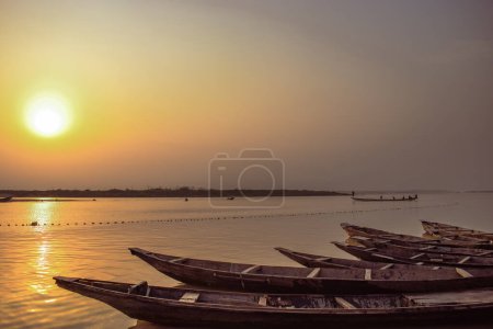 Téléchargez les photos : Delta State, Nigeria - 9 décembre 2021 : Canoës fabriqués localement au bord d'une rivière sur le fleuve Niger compensant un magnifique lever de soleil. Bateaux de pêche au bord d'une rivière. Réflexion sur une surface d'eau. - en image libre de droit