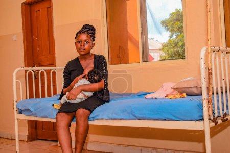 Foto de Jos, Nigeria - 12 de mayo de 2021: Madre africana con su hijo sentado en una cama de hospital. Mamá amamantando bebé. Mujer y niño recién nacido en el hospital - Imagen libre de derechos