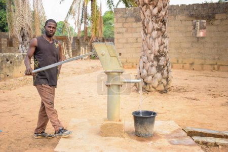 Foto de Karara, Nasarawa State - 5 de mayo de 2021: El hombre africano bombea agua de una bomba de mano Borewell bajo el sol en una comunidad rural - Imagen libre de derechos