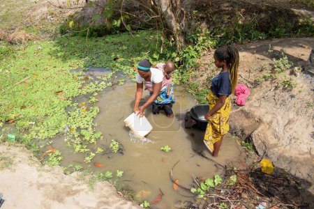 Foto de Karara, Estado de Nasarawa - 5 de mayo de 2021: Mujer africana con su bebé atrapando agua contaminada e inmunda de un estanque o arroyo para su consumo diario - Imagen libre de derechos