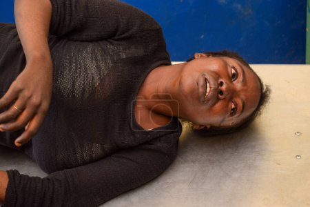 Foto de Lagos, Nigeria - 6 de septiembre de 2021: Dama Africana Vestida de Negro con Dolores Graves que Viene en Rueda para una Operación de Emergencia. Lágrimas debido a dolores abdominales. Apendicitis rota - Imagen libre de derechos