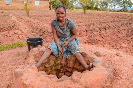 Téléchargez les photos : Jos East, État du Plateau - 12 septembre 2021 : Une adolescente africaine récupère l'eau d'un puits creusé à la main dans une communauté rurale. Eau boueuse brute non traitée - en image libre de droit