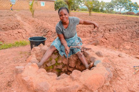 Foto de Jos East, Plateau State - 12 de septiembre de 2021: Adolescente africana cogiendo agua de un pozo abierto excavado a mano en una comunidad rural. Agua cruda sin tratar Muddy - Imagen libre de derechos