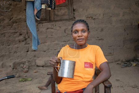Téléchargez les photos : Opialu, État de Benue - mars 6, 2021 : African Lady Wearing an Orange T-Shirt Drinking Water from a Stainless Cup outside her Home - en image libre de droit