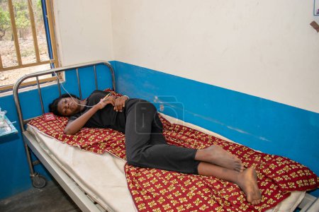 Téléchargez les photos : Opialu, État de Benue - mars 6, 2021 : Une adolescente africaine portant du noir est allongée sur un lit dans un centre de soins de santé primaires alors qu'elle reçoit un traitement. - en image libre de droit