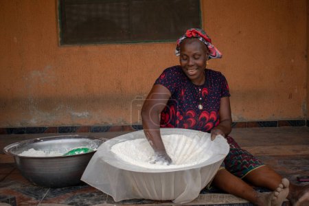 Foto de Opialu, Estado de Benue, Nigeria - 6 de marzo de 2021: Mujer africana de mediana edad sonriente trabajando y sentada en el suelo mientras tamiza la harina de mandioca del proceso (Garri) - Imagen libre de derechos