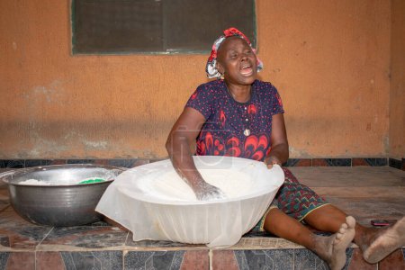 Téléchargez les photos : Opialu, État de Benue, Nigéria - 6 mars 2021 : Femme africaine souriante du Moyen Âge travaillant et s'asseyant sur le sol tout en tamisant le processus Farine de manioc (Garri) - en image libre de droit