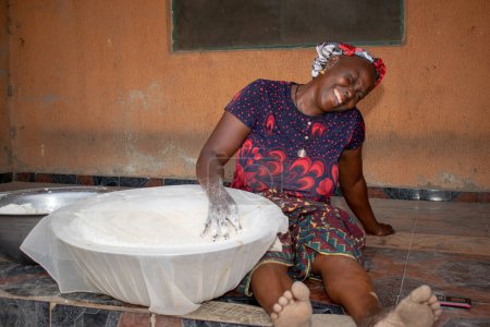 Foto de Opialu, Estado de Benue, Nigeria - 6 de marzo de 2021: Mujer africana de mediana edad sonriente trabajando y sentada en el suelo mientras tamiza la harina de mandioca del proceso (Garri) - Imagen libre de derechos