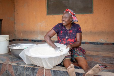 Téléchargez les photos : Opialu, État de Benue, Nigéria - 6 mars 2021 : Femme africaine souriante du Moyen Âge travaillant et s'asseyant sur le sol pendant le processus de tamisage Farine de manioc - en image libre de droit