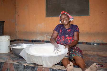 Photo for Opialu, Benue State, Nigeria - March 6, 2021: Smiling Middle Aged African Woman Working and Sitting on the Floor while Sieving Process Cassava Flour (Garri) - Royalty Free Image